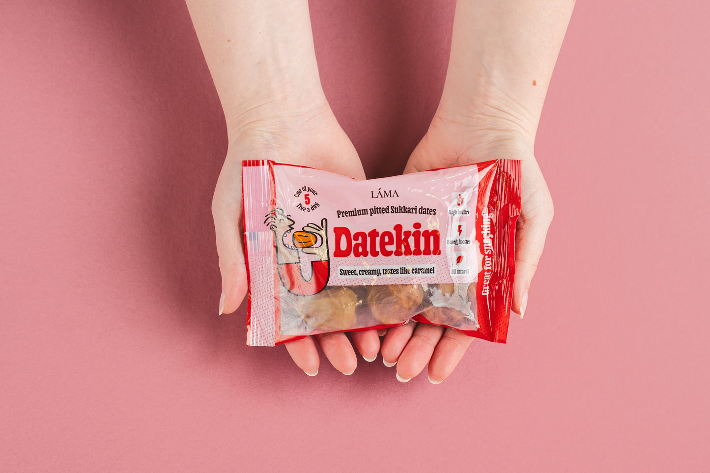 Hands hold a red packet of Datekin Premium Pitted Sukkari Dates against a pink background, showcasing the natural sweetness of healthy dates with illustrations and text including sweet, creamy, gooey, rich caramel.