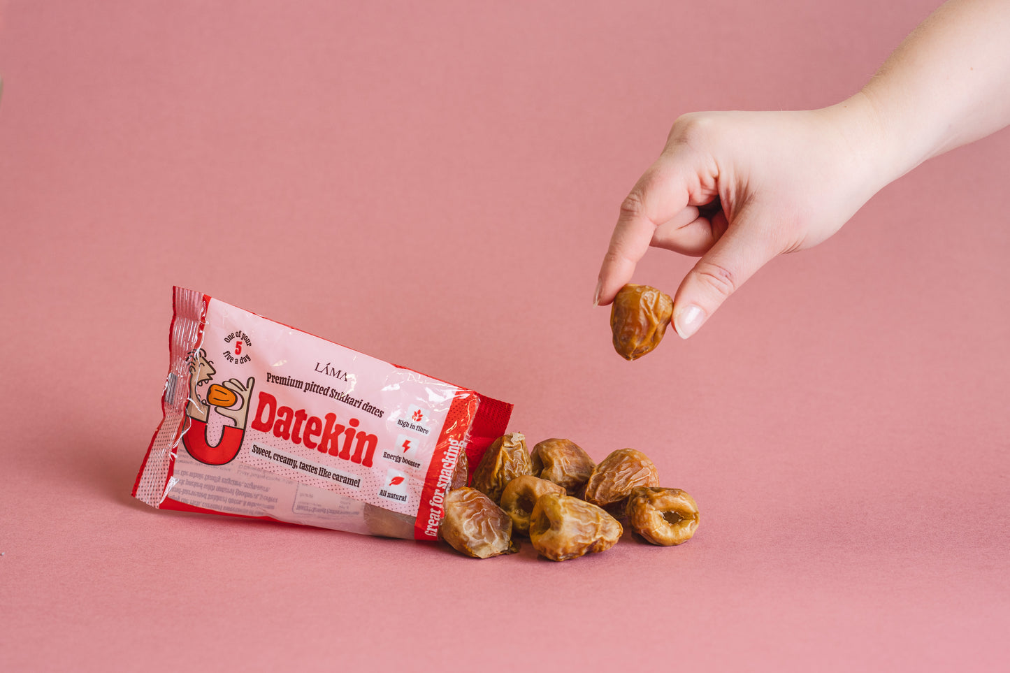 A hand picks up a date from a pile of loose Pitted Sukkari Dates on a pink surface. Beside them is the Datekin Premium Pitted Sukkari Dates red and white packaging, offering the perfect healthy snack thats always shelf ready.