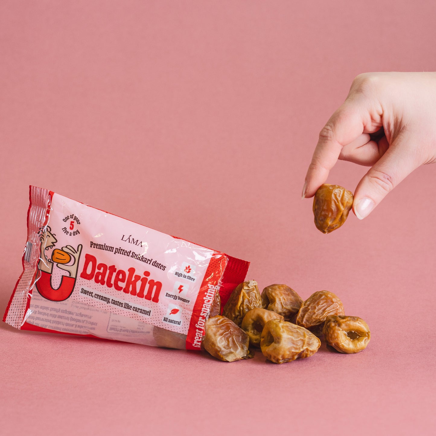 A hand picks up a date from a pile of loose Pitted Sukkari Dates on a pink surface. Beside them is the Datekin Premium Pitted Sukkari Dates red and white packaging, offering the perfect healthy snack thats always shelf ready.